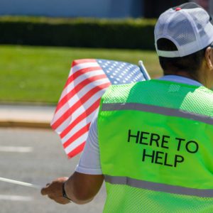 A person standing by a street wearing a neon vest reading "Here to Help."